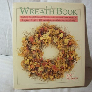 May include: The Wreath Book, a square book with a cream-colored cover, features a vibrant wreath made of dried flowers and foliage. The title is in green, with text below in red. The author's name is at the bottom.