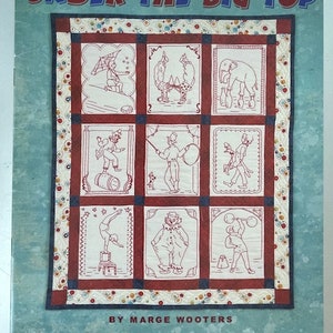 Op de afbeelding: Een quiltpatroonboek getiteld "Under the Big Top" met redwork-ontwerpen. De omslag heeft een blauwe achtergrond met een quiltblokontwerp met circus-thema-illustraties. Het boek is van Marge Wooters en uitgegeven door Indygo Junction, Inc.
