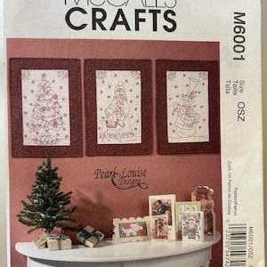 May include: McCall's Crafts sewing pattern M6001. The pattern features three framed Christmas designs and other holiday-themed decorations. A small Christmas tree and gift boxes are on a white table. The text "Pearl Louise Designs" is visible.
