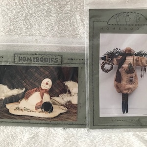 May include: Two Homebodies sewing patterns. The first pattern is for a snowman with a red and white scarf and a black hat. The second pattern is for a woman with a brown dress and a green wreath. The text on the patterns reads "Homebodies".