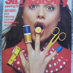 Puede incluir: Una portada de libro de costura vintage Simplicity con una mujer con una camisa de lunares rojos, blancos y azules. La mujer sostiene una regla de costura amarilla, un carrete de hilo y unas tijeras amarillas. El texto de la portada dice "Simplicity sewing book REVISED plus tailoring crochet embroidery and more".