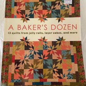 Puede incluir: Un libro titulado "A Baker's Dozen" con un patrón de colcha colorido en la portada. El libro es de That Patchwork Place y presenta 13 colchas hechas con rollos de jalea, pasteles en capas y más.
