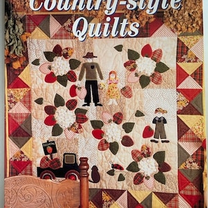 May include: A book cover featuring a patchwork quilt with a country-style theme. The quilt features a red truck, a scarecrow, a girl, and strawberries. The book title is "Fabulous Country-style Quilts" and the text "20 Inspirational projects Pattern sheet included" is at the bottom of the cover.