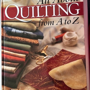 Könnte beinhalten: Ein Buch mit dem Titel "All About Quilting from A to Z" mit einem rot-weißen Einband. Das Bild zeigt Stapel farbiger Stoffe, eine Schere, Faden und andere Quilt-Zubehörteile. Das Buch wird von Quilter's Newsletter Magazine, Quiltmaker Magazine und C&T Publishing herausgegeben.