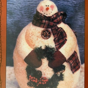 Può includere: Un pupazzo di neve bianco con il naso rosso e gli occhi neri, che indossa una sciarpa a quadri e tiene una ghirlanda verde con bacche rosse.