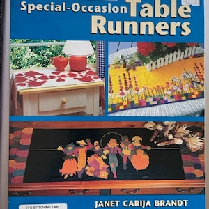 May include: A book titled "Special-Occasion Table Runners" by Janet Carija Brandt. The book cover features three different table runner designs: a red heart design, a yellow and green floral design, and a black and orange scarecrow design.