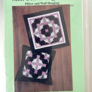 May include: A pattern for a mini Denver log cabin quilt, featuring a black and pink design with white accents. The pattern is for a pillow and wall hanging. The pattern is designed by Lola Hendrickson and is available from the Quilt Company in Blue Earth, Minnesota.