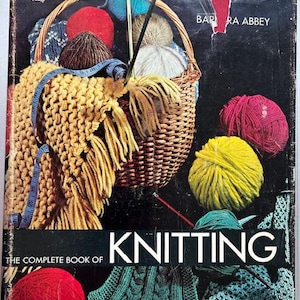 Può includere: Una copertina di libro vintage intitolata "Knitting: The Complete Book of Knitting" con un cestino di filato e ferri da maglia. La copertina presenta una sciarpa gialla lavorata a maglia, gomitoli di lana rossi e gialli e un metro a nastro blu.