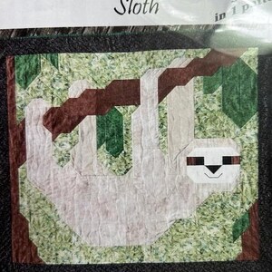 Peut inclure: Un patron de quilt avec un motif de paresseux. Le paresseux est blanc avec un visage noir et marron, suspendu à une branche marron. Le fond est vert avec un motif de feuilles. Le texte "Sloth" est au-dessus de l'image.