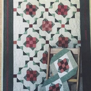 Peut inclure: Une courtepointe avec un fond blanc et un motif de carrés rouges et verts. La courtepointe est pliée et drapée sur une chaise en bois.