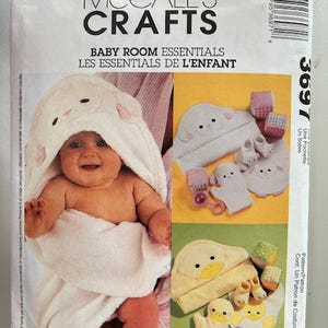 Op de afbeelding: Een McCall's Crafts patroon voor babykamer essentials. Het patroon toont een baby gewikkeld in een witte badhanddoek met berenoren. Het patroon toont ook voorbeelden van een badhanddoek met capuchon, babyslofjes en washandjes.