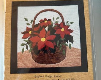 Peut inclure: Un kit de bricolage intitulé "Poinsettia Basket" avec un motif de tissu de poinsettias rouges dans un panier marron. Le kit comprend des patrons thermocollants. L'emballage a des bordures décoratives et le texte "England Design Studios".