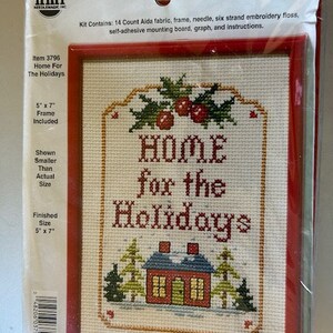 Op de afbeelding: Cross stitch-set met een rode lijst, met een witte achtergrond met de tekst "Home for the Holidays" in rode en groene letters. Het ontwerp bevat een klein rood huis met witte versieringen en sneeuw op het dak, twee groene bomen en een tak met rode bessen.