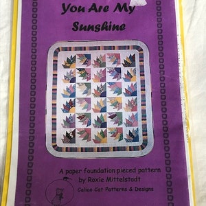 Peut inclure: Un modèle de patchwork en papier à fond uni intitulé "You Are My Sunshine" par Roxie Mittelstadt. Le modèle présente un motif de courtepointe avec un motif de soleil éclatant et provient de Calico Cat Patterns & Designs.