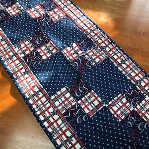 May include: A red, white, and blue quilted table runner with a patchwork design. The runner features a navy blue background with white polka dots and red and white plaid squares.