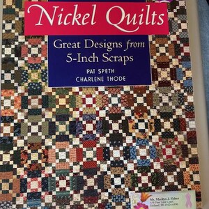 May include: A book titled "Nickel Quilts" with the subtitle "Great Designs from 5-Inch Scraps" by Pat Speth and Charlene Thode. The cover features a patchwork quilt design in various colors, including red, blue, green, and brown. A pink sticker with the number 2 is on the top right corner.