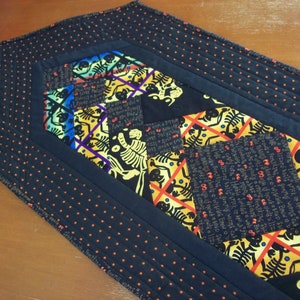 Peut inclure: Un chemin de table noir à pois orange et un motif patchwork coloré avec des squelettes et des crânes. Le chemin de table est matelassé et a une bordure noire.