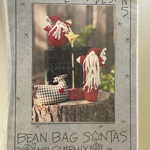 May include: A package featuring a photo of handmade bean bag Santas. The Santas are red with white accents and long white beards. One Santa is black and white checkered with a red heart. The text reads "On The Edge Designs" and "Bean Bag Santas and Company."