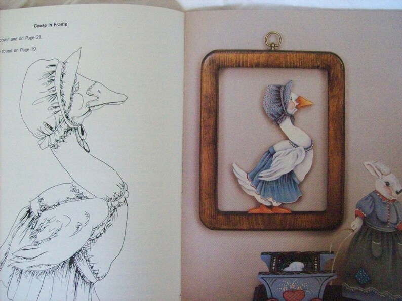May include: A black and white line drawing of a goose wearing a bonnet and dress. The goose is in a frame with a painted goose wearing a bonnet and dress. The painted goose is in a blue dress with white accents. The goose is holding a string attached to a painted rabbit wearing a blue dress with white accents.