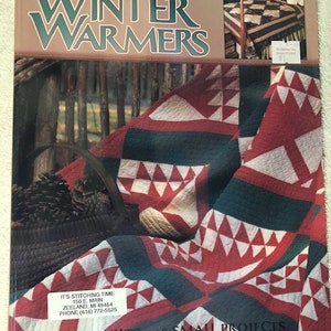May include: A book cover with the title "Winter Warmers" featuring a red, white, and green quilt with a basket of pine cones in the foreground. The book cover also includes the text "14 Quilts - 16 Small Projects" and the text "Quick Rotary Methods".