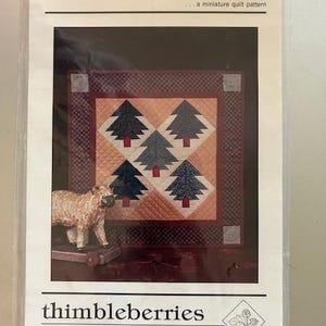 May include: A quilt pattern titled "Small Pines... a miniature quilt pattern" by Thimbleberries. The pattern features a quilt block with five stylized pine trees in shades of blue, green, and red on a peach background. The quilt block is surrounded by a dark red border with a white and black checkered pattern.