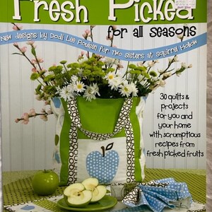 Op de afbeelding: Een boek getiteld "Fresh Picked for all seasons" met een groene en bruine tas met appelmotief, groene appels en een servet met een blauw-wit patroon. De omslag van het boek toont een bloemstuk en de tekst "New designs by Dodi Lee Poulsen for Two sisters at Squirrel Hollow".