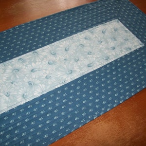 May include: A blue and white quilted table runner with a floral pattern. The runner has a blue border with a white floral pattern in the center.