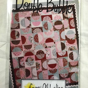 May include: Quilt pattern titled "Double Bubble" by Crazy Old Ladies. The pattern features a patchwork design with pink, brown, and blue circles and half-circles. The pattern is $9.25.