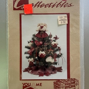May include: A craft pattern for a country-style Christmas tree with a doll on top, ornaments, and a red and white fabric base. The pattern is from You & Me Patterns Inc. and is titled "Country Needleworks".