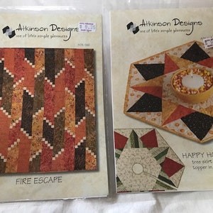 May include: Two quilting patterns, one titled "Fire Escape" with a geometric pattern in shades of orange, brown, and white, and the other titled "Happy Holidays" with a star-shaped design in black, orange, and white, with a candle holder in the centre.