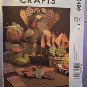 May include: A sewing pattern for a Thanksgiving turkey, pumpkin, and place card holders. The pattern includes instructions for making a turkey with a plaid body, a pumpkin with the words "Give Thanks", and place card holders with leaves. The pattern is from McCall's Crafts and is size OSZ.