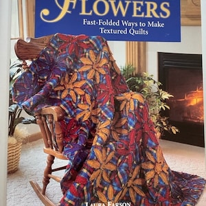 Może przedstawiać: Książka zatytułowana "Ragged-Edge Flowers" z podtytułem "Fast-Folded Ways to Make Textured Quilts". Kolorowa kołdra z kwiatowymi wzorami jest narzucona na drewniany fotel bujany. Kołdra ma odcienie czerwieni, błękitu i pomarańczy.