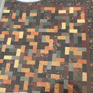 以下が含まれることがあります： 「GRAHAM CRACKERS」と「Pacific Patchwork」の文字がパッケージに書かれたキルトキット。キルトは、黒、茶色、オレンジ、緑、タンの色合いの幾何学模様が特徴です。