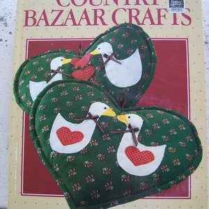 Puede incluir: Un libro titulado "Country Bazaar Crafts" con agarraderas en forma de corazón con patos blancos con detalles de corazón rojo. Las agarraderas son verdes con un estampado floral. El libro está sobre una superficie beige.