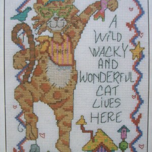 May include: A cross-stitch pattern featuring a cartoon cat wearing glasses and a striped shirt. The cat is holding a fish and standing in front of a house. The text "A wild wacky and wonderful cat lives here" is written on the right side of the image.