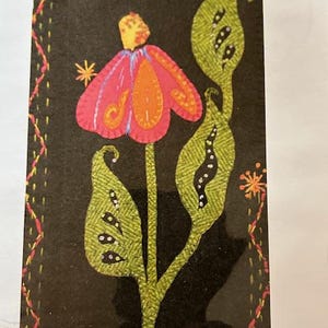 May include: A felt applique featuring a stylized flower with a red and orange blossom, green leaves, and a yellow center. The design is set against a black background with a decorative stitched border and star accents. The dimensions are 6 x 10 inches.