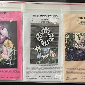 May include: Three vintage craft patterns for making a cow wreath, a pair of love knot dolls, and a pigtail doll. The patterns are printed on pink, beige, and white paper with illustrations and instructions. The patterns are titled "Moo-ving 'Round", "Love Knots", and "Pig Tails".