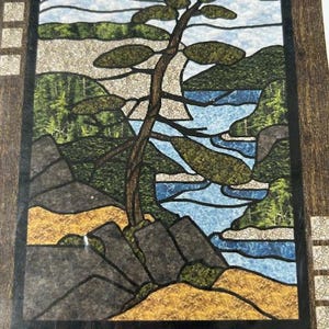 May include: Stained glass art piece depicting a landscape scene. The artwork features a tree with green foliage, a blue river, and rocky terrain. The piece is framed in a dark wood border, with a detailed design.
