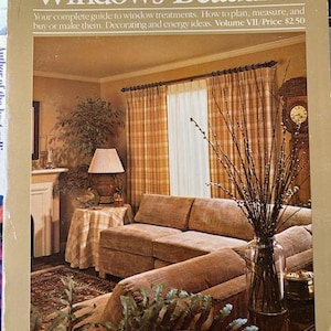 May include: A book cover titled "Windows Beautiful" with text about window treatments, decorating, and energy ideas. The image shows a living room with a brown sectional sofa, plaid curtains, and a table lamp.