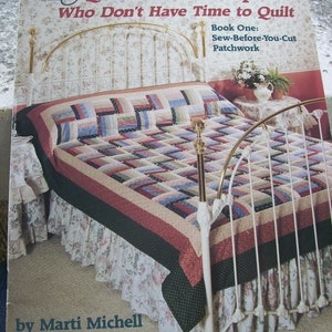 May include: Book cover for "Quilting for People Who Don't Have Time to Quilt" by Marti Michell. The cover features a white bed with a red, blue, and white patchwork quilt. The title of the book is in large, bold letters.