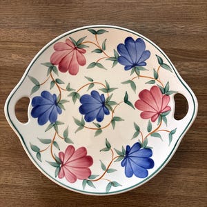 May include: A round ceramic serving platter with two integrated side handles. The off-white surface features a hand-painted pattern of alternating pink and blue scalloped flower blossoms connected by thin orange vines and small green leaves.
