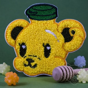 Op de afbeelding: Gele geborduurde patch van een beer-vormige honingfles met een groen deksel. De beer heeft zwarte ogen en een zwarte neus.