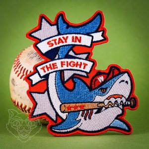 May include: Embroidered patch featuring a cartoon shark wearing a baseball cap with a 'W' on it, holding a baseball bat in its mouth. The shark is blue and white with red accents. The patch has a red background with a white banner that says 'Stay in the Fight'.