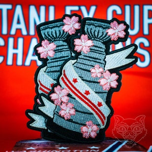 May include: Embroidered patches featuring a stylized hockey trophy design, adorned with pink cherry blossoms and a banner with red stripes and stars. The patches have a black border and are set against a blurred background.