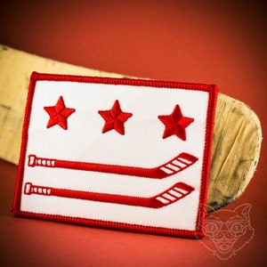 May include: White embroidered patch with a red border. The patch features three red stars and three red hockey sticks, representing the Washington D.C. flag.