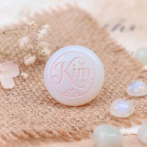 May include: A round, white, opaque cabochon featuring the name Kim engraved in a cursive script with a small lotus flower icon below the text. The surface is smooth and polished, displaying a soft, milky appearance with subtle light reflections.