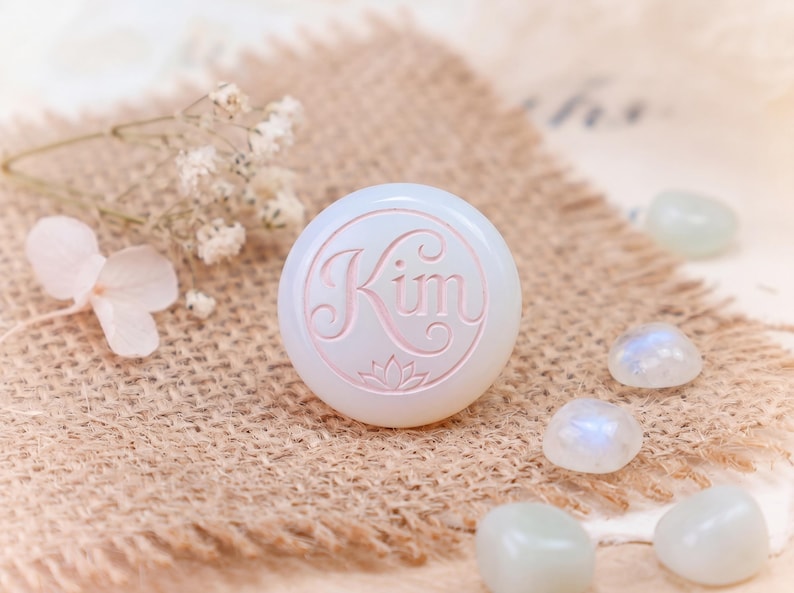 May include: A round, white, opaque cabochon featuring the name Kim engraved in a cursive script with a small lotus flower icon below the text. The surface is smooth and polished, displaying a soft, milky appearance with subtle light reflections.