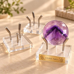 May include: A polished purple amethyst sphere with a dark chevron pattern resting on a clear acrylic stand with silver metal prongs. The base features a gold-toned rectangular plaque with the text Crystal Aura and leaf illustrations.