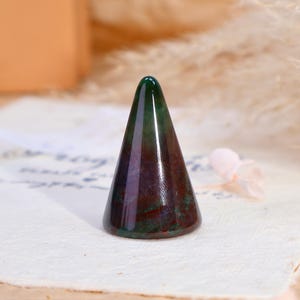 May include: A polished stone ring holder shaped like a cone, featuring a smooth surface with deep green, dark red, and purple marbled color variations throughout the mineral structure, standing upright on a flat circular base.