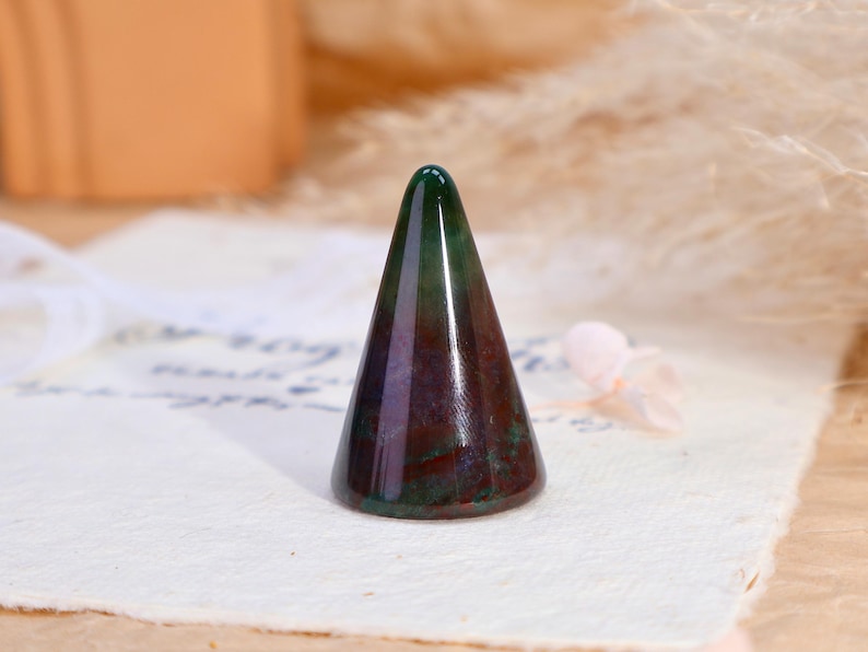 May include: A polished stone ring holder shaped like a cone, featuring a smooth surface with deep green, dark red, and purple marbled color variations throughout the mineral structure, standing upright on a flat circular base.